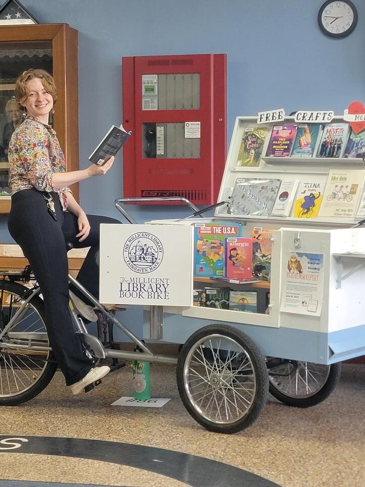 Youth Services Librarian Allie perches with a book in hand atop the Book Bike seat. The bike, parked at the Middle School Vendor fair is a front-loaded cargo bike, open and overflowing with books on display.